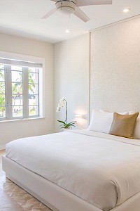 The image shows a bright, modern bedroom with a large bed, neutral colors, a ceiling fan, and a window with palm trees visible outside.