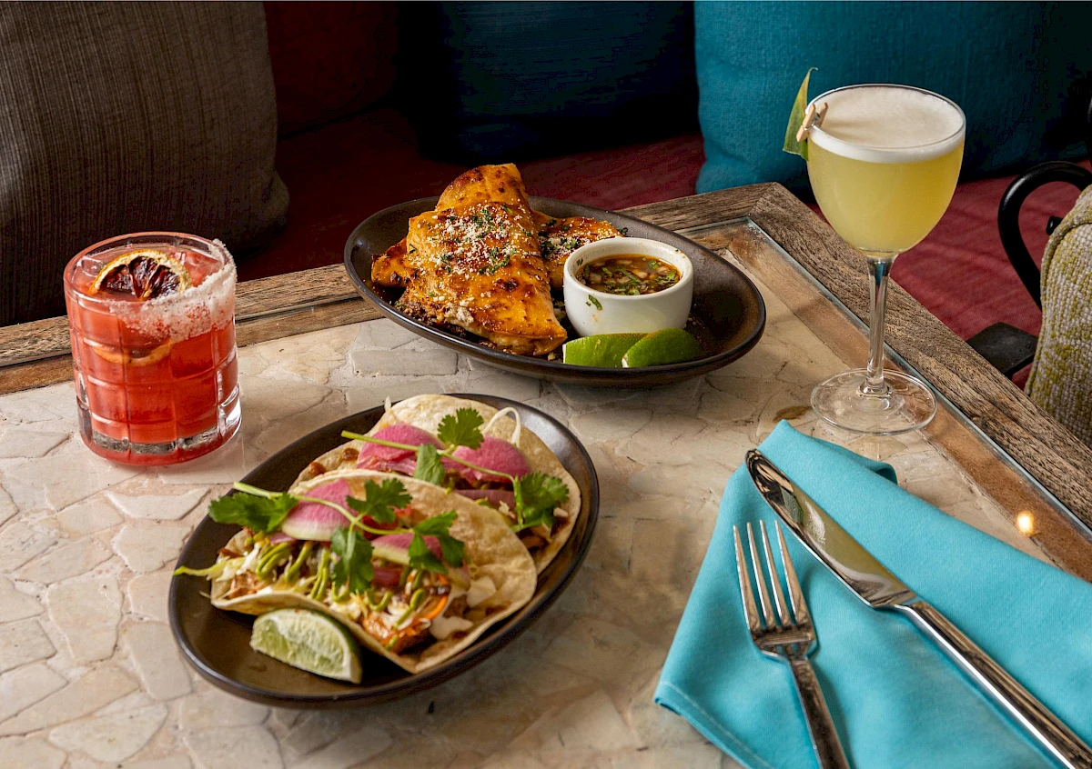 The image shows a table with Mexican dishes, cocktails, a fork, a knife, a blue napkin, and colorful cushions in the background.