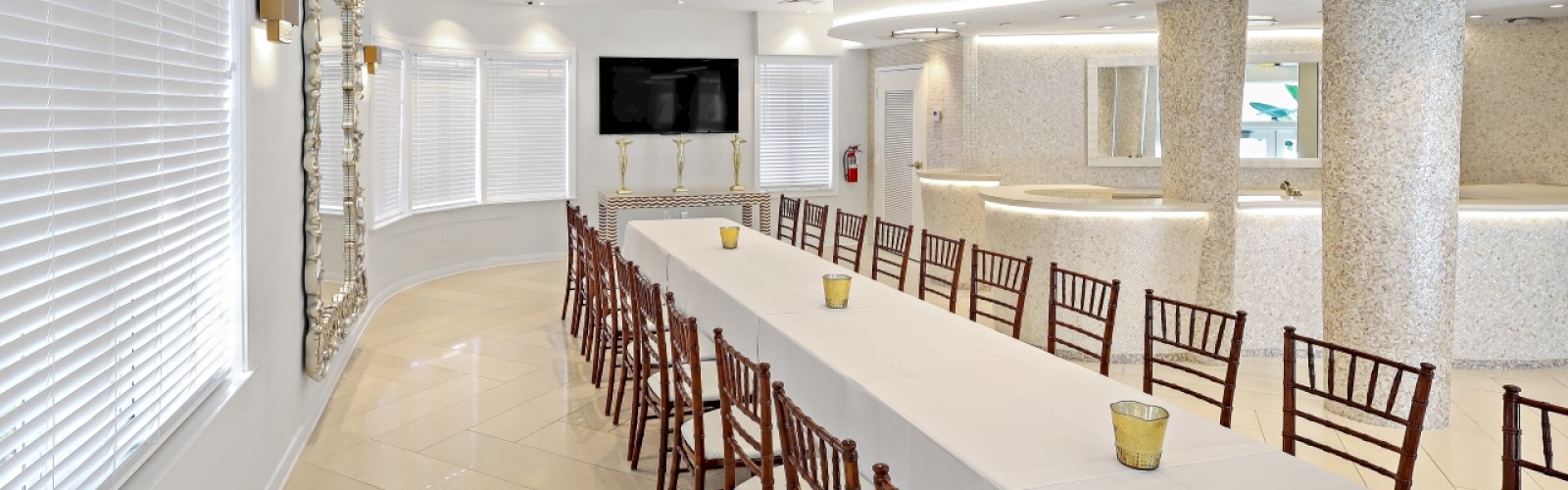 A long banquet table with white tablecloth, brown chairs, and small candles is set up in a modern, bright room with white walls and large windows.