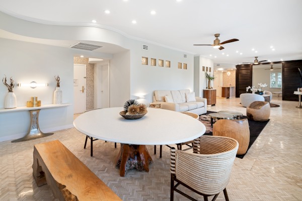 A modern living space with neutral tones, featuring a round table, seating area, decorative items, and ceiling fans on a tiled floor.