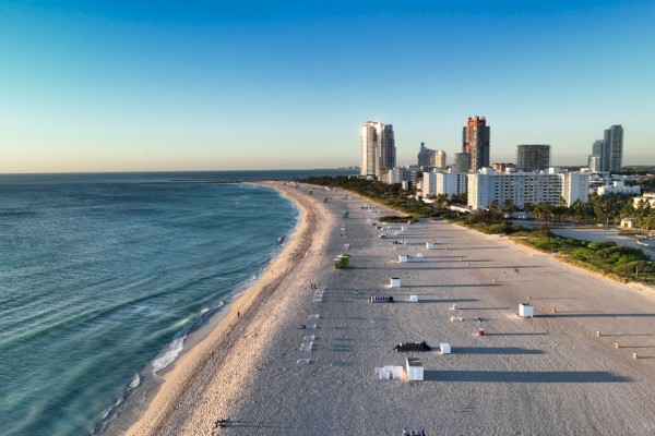 A beach with calm waves, white sand, and a city skyline in the background under a clear blue sky. Beach chairs and cabanas are arranged.
