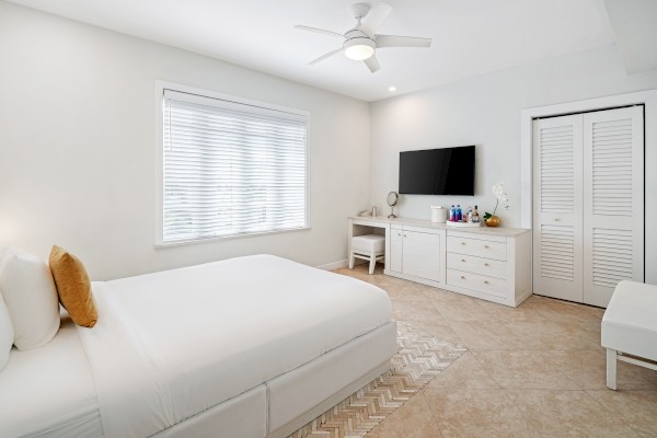 A bright bedroom with a large bed, TV, dresser, and window. It has tile flooring, a ceiling fan, and a desk with a chair.