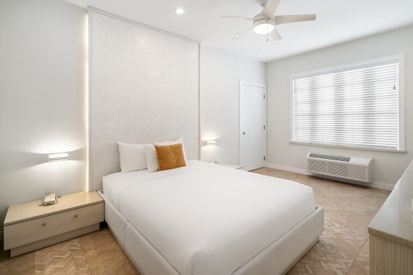 A modern bedroom with a double bed, light wood furniture, ceiling fan, and large window with blinds in a bright, minimalist setting.