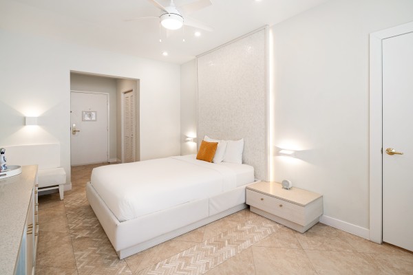 A modern, minimalist bedroom with a white bed, side table, wall art, and ceiling fan. Neutral tones and sleek design create a calming vibe.