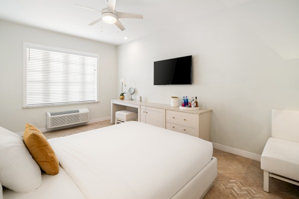 A minimalist bedroom with a white bed, TV, dresser, chair, and window with blinds. A ceiling fan and desk items complete the look.