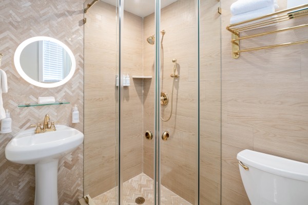 The image shows a modern bathroom with a glass shower enclosure, pedestal sink, and toilet. Elegant fixtures and neutral tiles create a sleek finish.