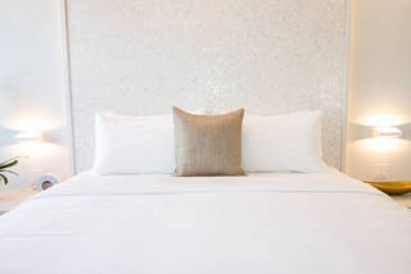 This image shows a neatly made bed with white bedding, flanked by modern wall-mounted lamps, and features a single beige throw pillow in the center.