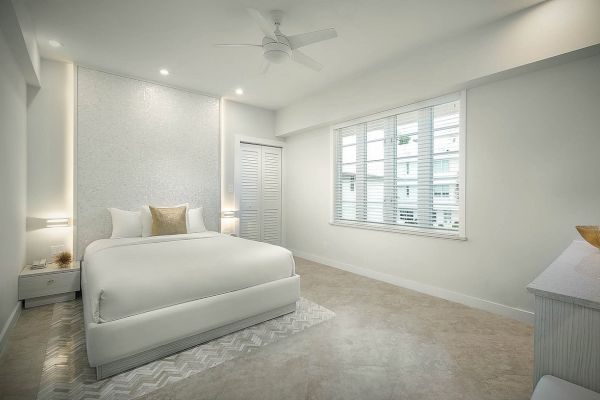 A minimalistic bedroom with a white bed, wooden floor, ceiling fan, and a large window with blinds, creating a bright and airy ambiance.