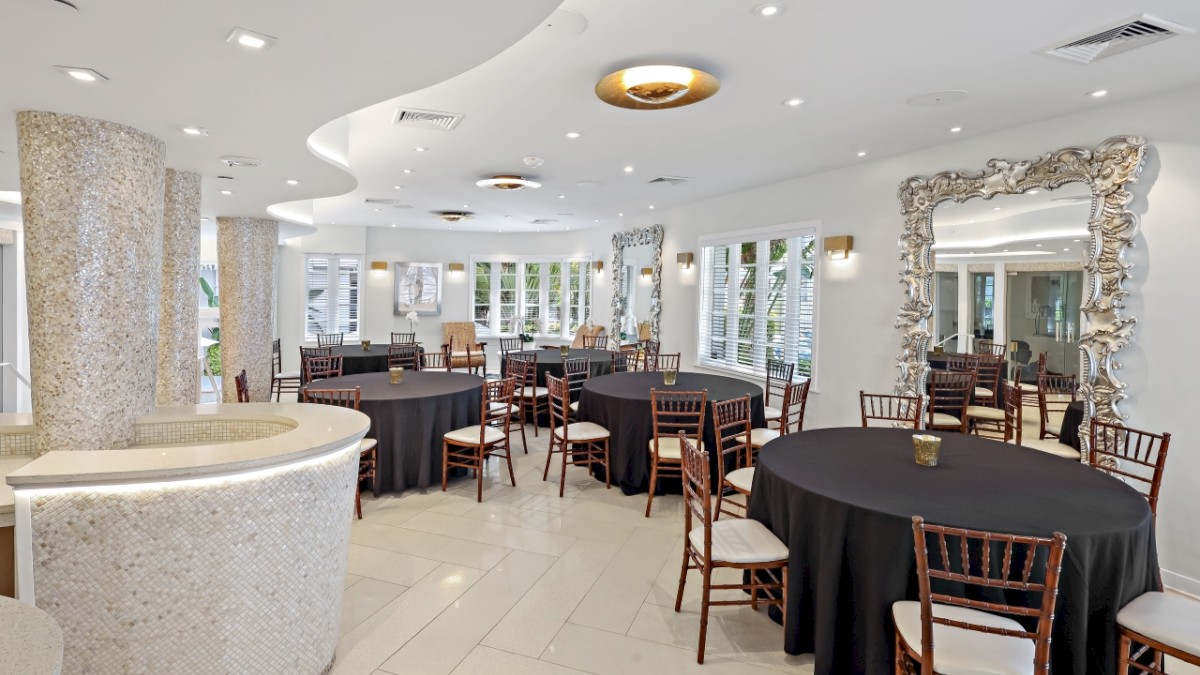 The image shows a modern, elegantly decorated dining area with round tables covered in black tablecloths and wooden chairs, large mirrors, and ample lighting.