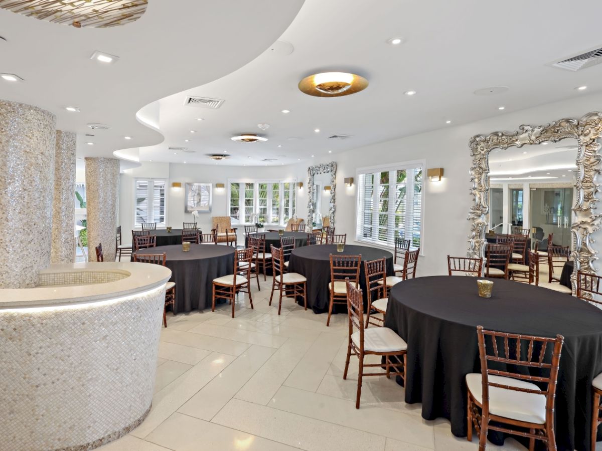 The image shows a modern, elegantly decorated dining area with round tables covered in black tablecloths and wooden chairs, large mirrors, and ample lighting.