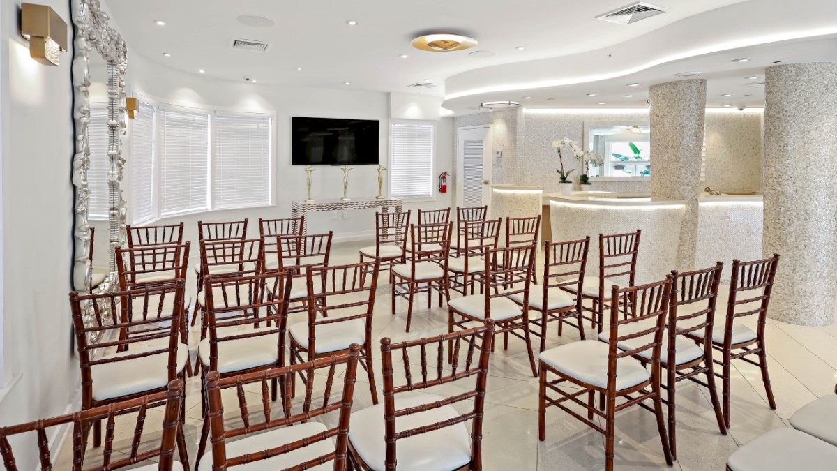 The image shows a bright, modern conference or event room with rows of wooden chairs arranged facing a large screen on the wall.