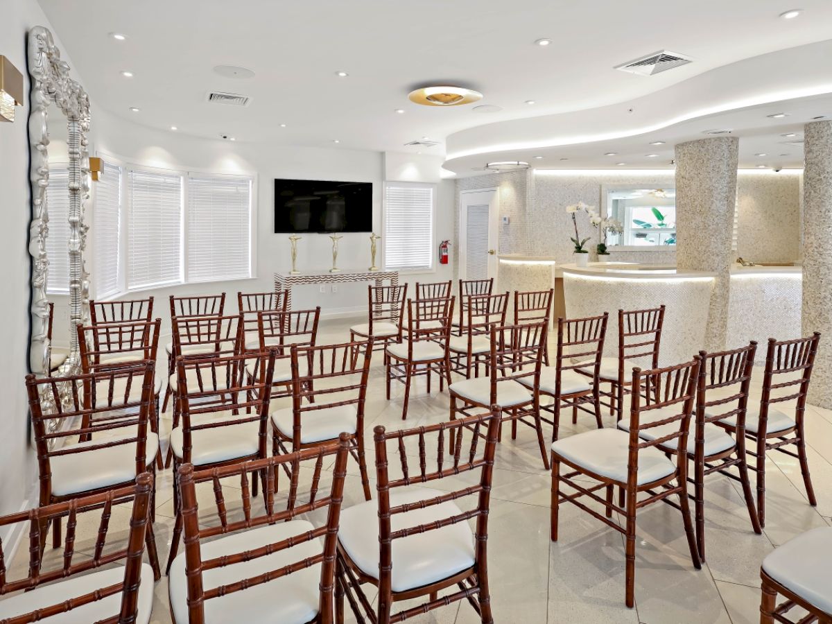 The image shows a bright, modern conference or event room with rows of wooden chairs arranged facing a large screen on the wall.