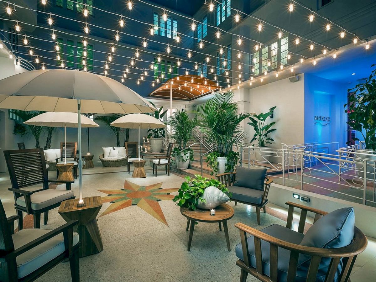 An outdoor patio with string lights, umbrellas, and wooden seating arranged around tables. The area is decorated with potted plants, creating a cozy ambiance.