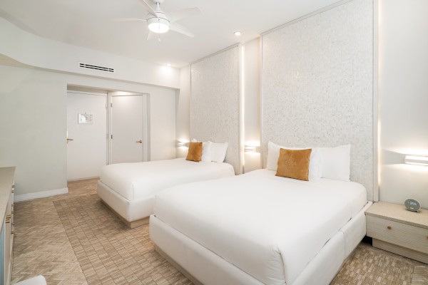 The image shows a modern hotel room with two neatly made beds, a neutral color palette, and soft lighting, creating a cozy atmosphere.