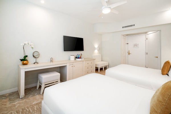 A hotel room with two beds, a TV, a desk with a mirror, a vanity stool, and a door. Neutral colors and a minimalist design complete the scene.