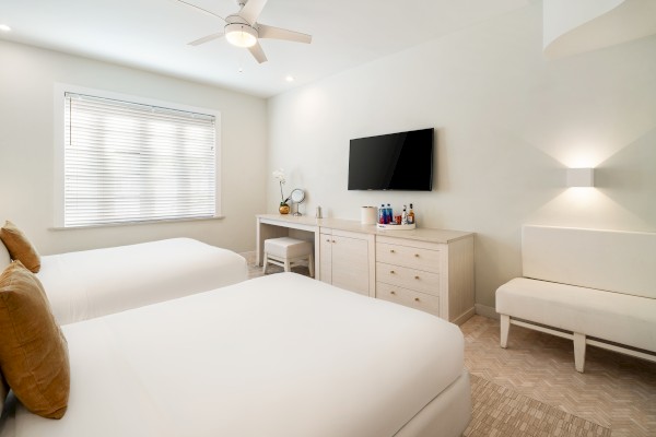 A minimalist hotel room with two beds, a TV, a dresser, a desk with a mirror, and a bench, decorated in neutral tones.