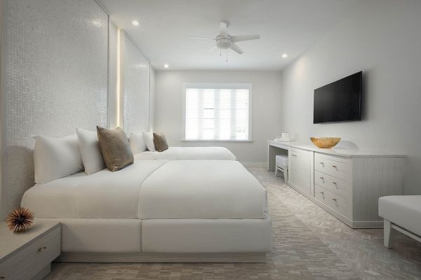 This image shows a modern, minimalist bedroom with two beds, a wall-mounted TV, a dresser, a ceiling fan, and neutral-toned decor, ending the sentence.