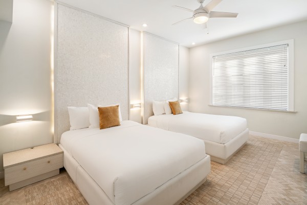 Modern bedroom with two white beds, accent pillows, nightstands, a ceiling fan, and a window with blinds. Light and airy decor.