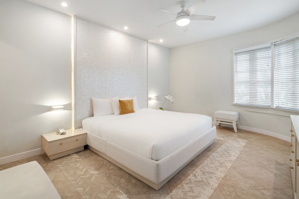 A modern bedroom with a large bed, two nightstands, a ceiling fan, and a window with blinds. Neutral tones and minimal decor complete the look.