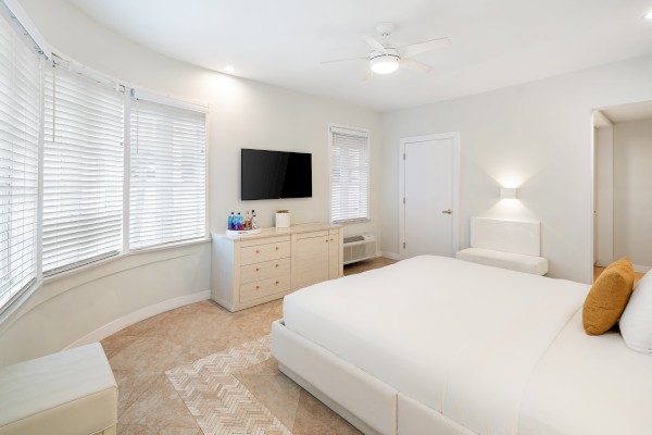 The image shows a bright bedroom with a large bed, dresser with a TV, windows with blinds, and a small seating area.