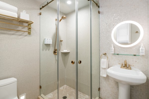 A modern bathroom with a glass shower, pedestal sink with a circular mirror, toilet, and towel rack on a light-colored tiled wall.