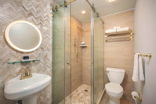 A bathroom with a sink, circular mirror, toilet, glass-enclosed shower, towel rack with towels, and wall-mounted toilet paper holder.