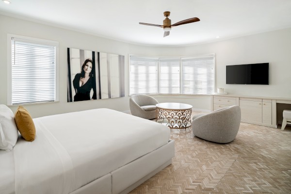 The image shows a modern bedroom with a large bed, two chairs, a round table, a TV, and a wall art piece.