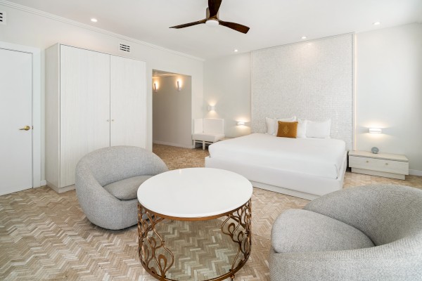 A modern bedroom features a large bed, two gray chairs, a round table, and a ceiling fan, all in a neutral-toned palette.