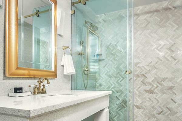 The image features a modern bathroom with a white countertop, gold-framed mirror, glass shower enclosure, and a wall with a herringbone tile pattern.