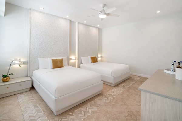 A modern bedroom with two double beds, white decor, a ceiling fan, bedside tables, and a console with drinks and a plant.