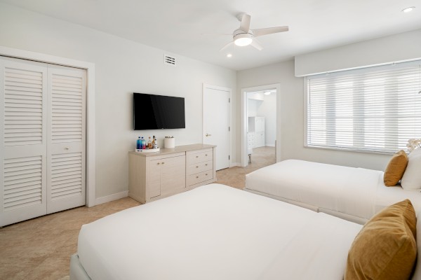 A bright bedroom with two beds, wall-mounted TV, dresser, and a large window with blinds. There's a fan on the ceiling.