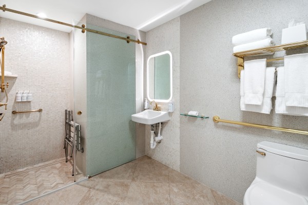 The image shows a modern bathroom with a shower, sink, toilet, and towel racks. It has gold accents and white towels on shelves.