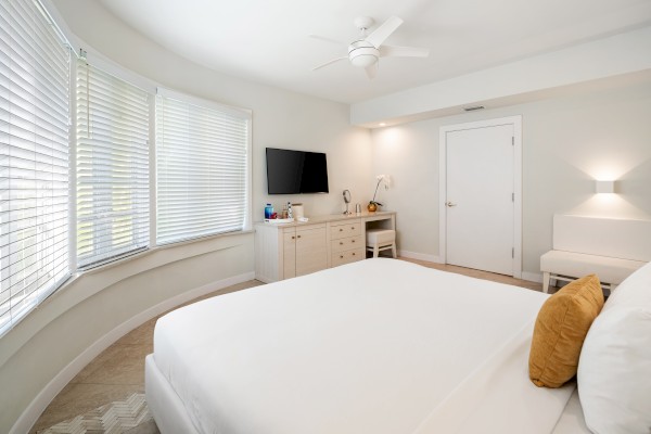 A bright bedroom features a bed with white linens, a mustard pillow, a TV, a dresser, and a ceiling fan, with large windows on the left.