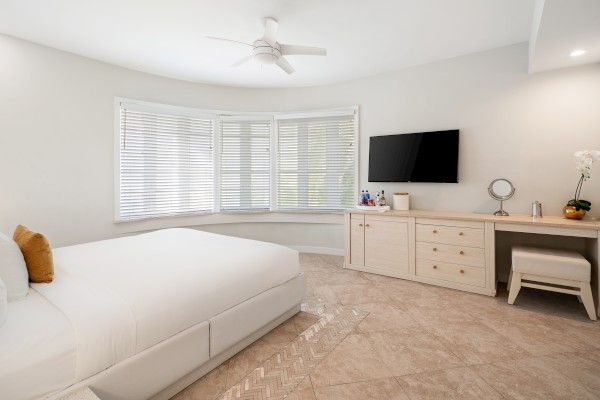 A modern bedroom with a large bed, dresser, flat-screen TV, and a window with blinds. Room features neutral tones and a ceiling fan.