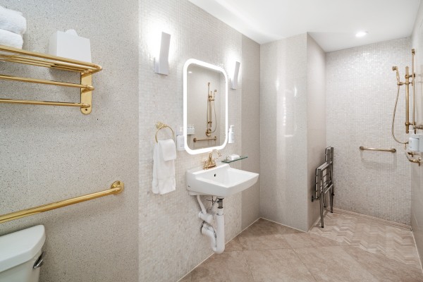 This image shows a modern bathroom with a sink, illuminated mirror, gold fixtures, and an open shower area.