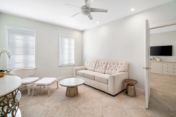 A modern living room with a beige sofa, round tables, chairs, and a ceiling fan. A doorway reveals a TV and dresser in the next room.