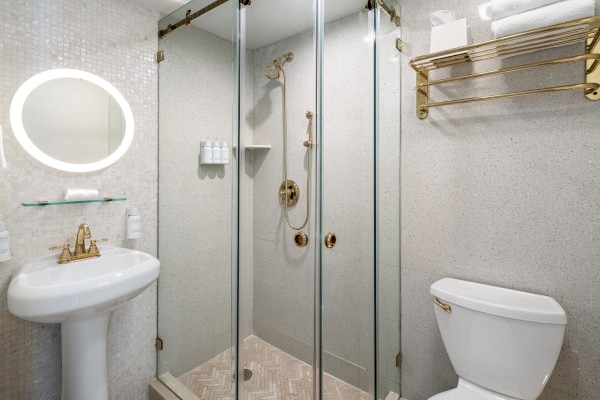 A modern bathroom with glass shower enclosure, pedestal sink with a round mirror, and a toilet. Towels and toiletries are neatly arranged.