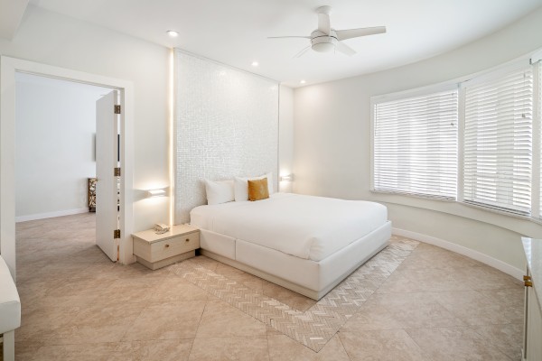 A bright, modern bedroom with a white bed, neutral decor, large windows with blinds, and a ceiling fan.