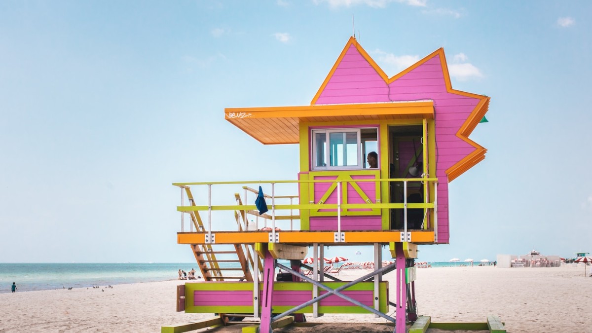 A vibrant, uniquely-shaped lifeguard tower in pink and orange stands on a sandy beach with the ocean and a few beachgoers in the background.