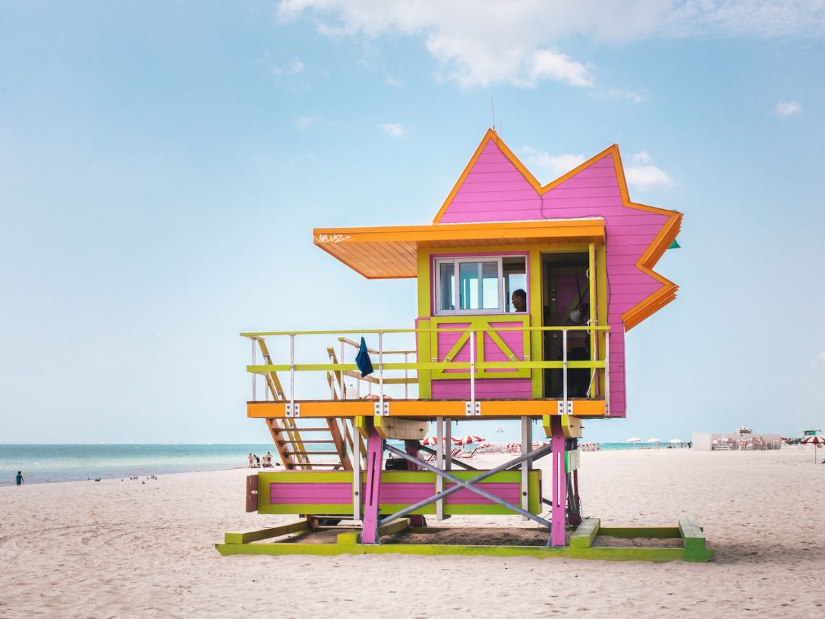 A vibrant, uniquely-shaped lifeguard tower in pink and orange stands on a sandy beach with the ocean and a few beachgoers in the background.