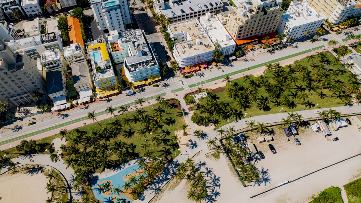 An aerial view shows a beachfront cityscape with buildings, palm trees, and beach amenities separated by a road.