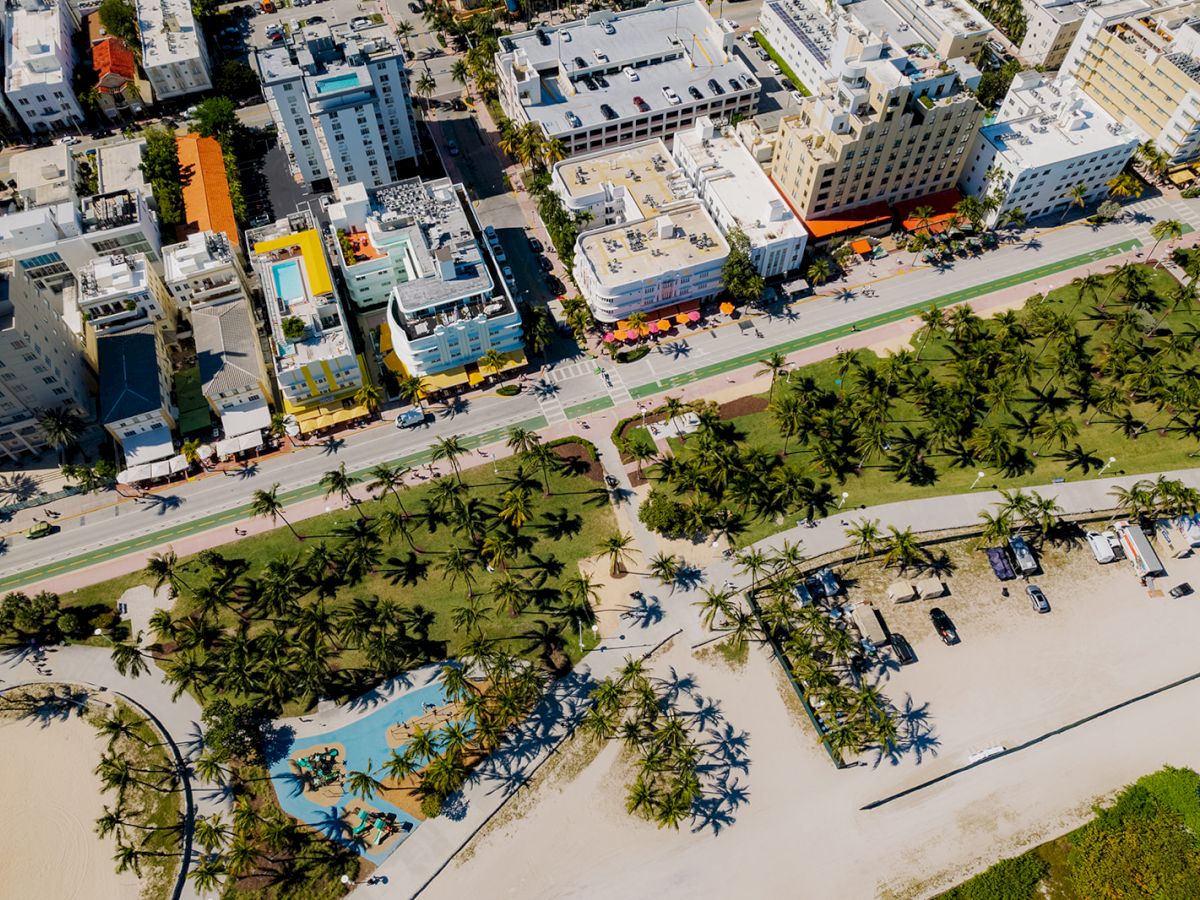 An aerial view shows a beachfront cityscape with buildings, palm trees, and beach amenities separated by a road.