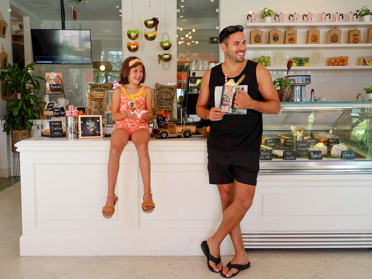 A man and a girl are in an ice cream shop. The girl is sitting on the counter, and the man is standing, both enjoying ice cream cones.