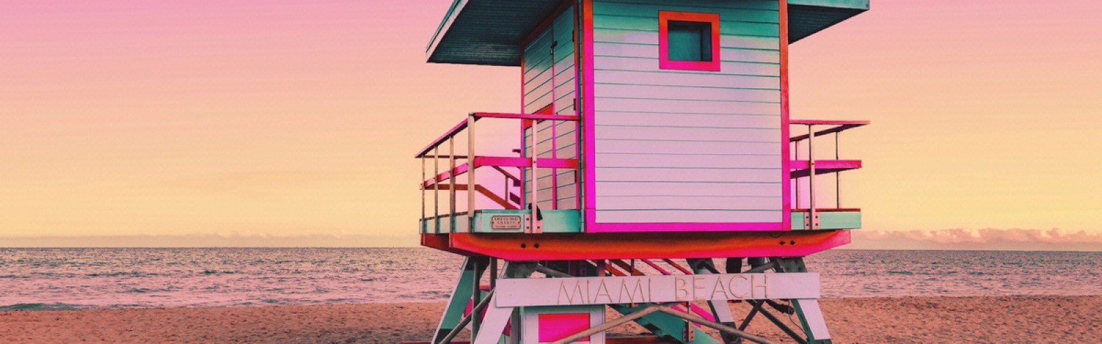 A brightly colored lifeguard tower on a sandy beach with a pink and yellow sky in the background.