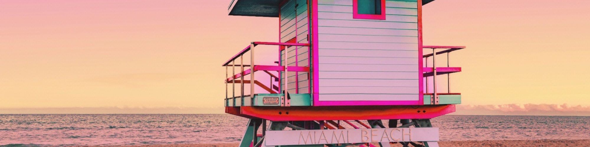 A brightly colored lifeguard tower on a sandy beach with a pink and yellow sky in the background.