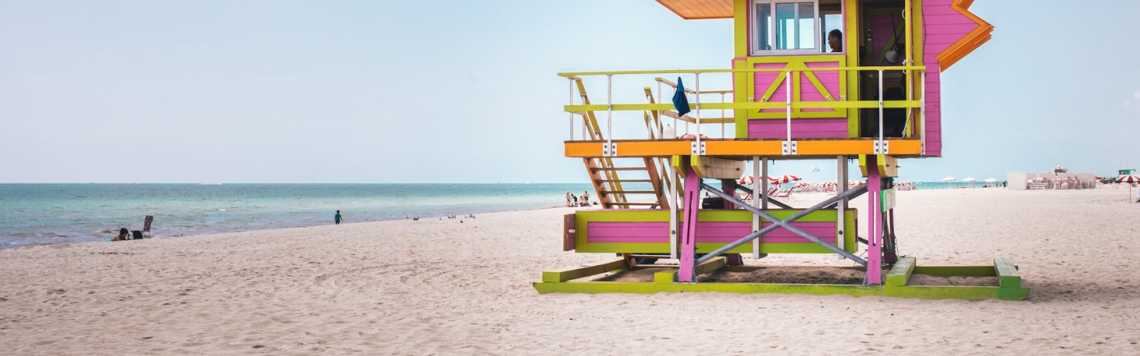 A colorful lifeguard tower stands on a sandy beach under a blue sky with scattered clouds and a few people in the distance.
