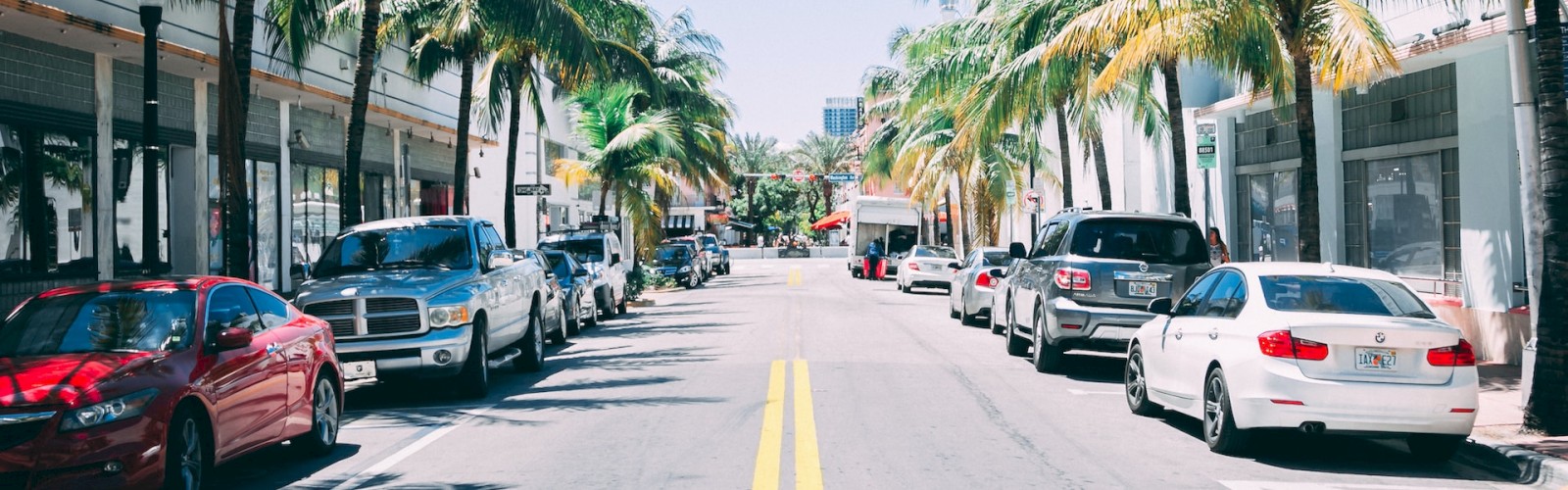 A sunny street lined with palm trees and parked cars on both sides, leading towards the horizon, suggesting a tropical or coastal city setting.
