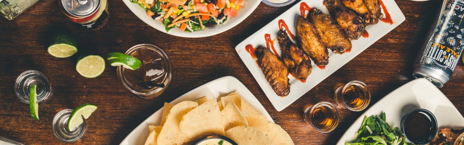An overhead view of a variety of dishes including wings, nachos, salad, drinks, burger, and sauces, arranged on a wooden table to share.