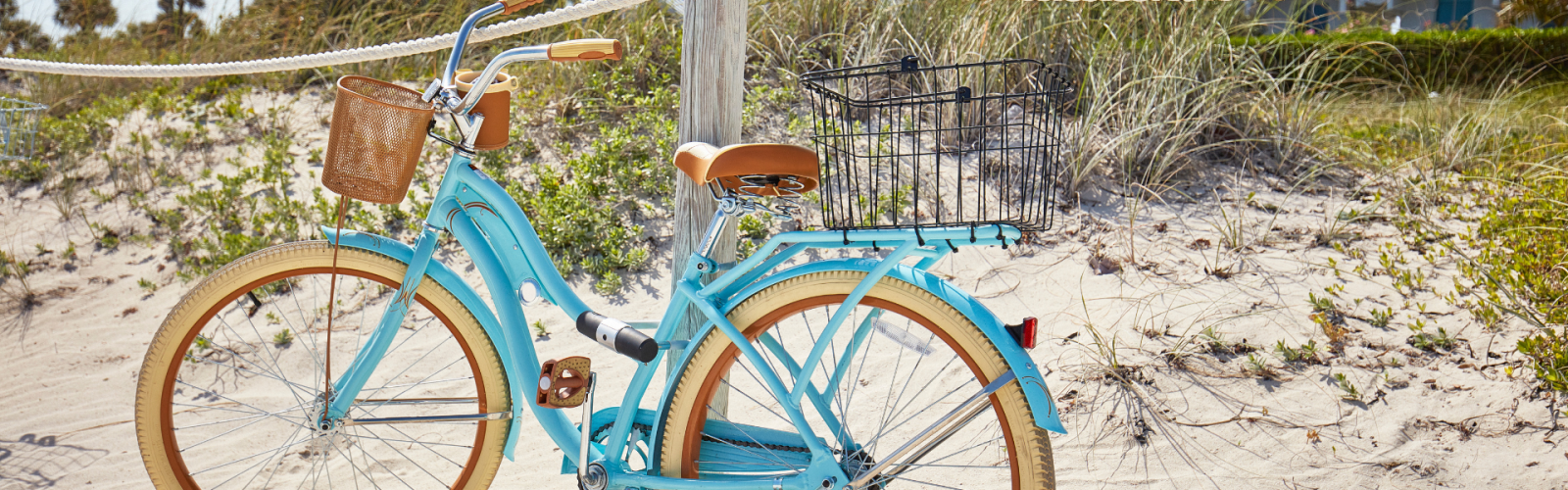 A light blue bicycle with a wicker basket is parked against a wooden post on a sandy beach. Palm trees and greenery are in the background.