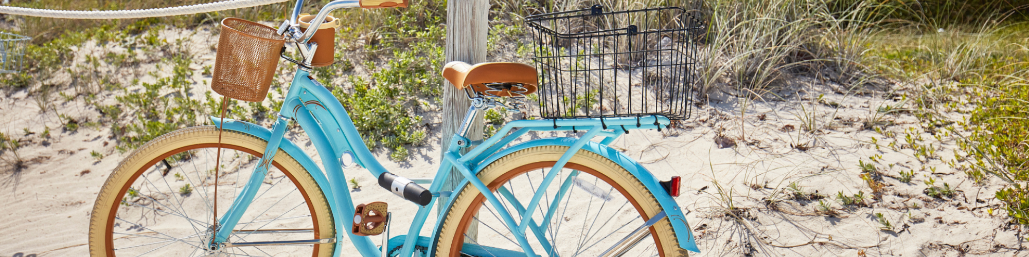 A light blue bicycle with a wicker basket is parked against a wooden post on a sandy beach. Palm trees and greenery are in the background.